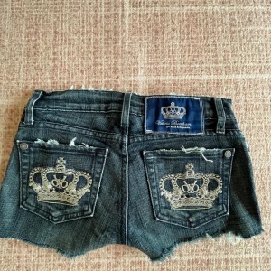 Mörkblå jeansshorts från Victoria Beckham - Super Snygga mörkblå jeansshorts från True Religion med fransiga kanter och broderade kronor på bakfickorna. Perfekta för en avslappnad stil med en touch av lyx. De har en knappgylf och klassisk femficksdesign.andvänd några gånger 