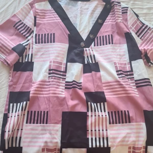 Mönstrad kortärmad blus med knappar - Säljer en färgglad blus med ett geometriskt mönster i rosa, svart och vitt. Blusen har en v-ringning med knappar och korta ärmar. Perfekt för en trendig look!