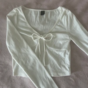 Vit ribbad topp från Shein - Säljer en söt vit ribbad topp från Shein med långärmad design och en knytrosett framtill. Toppen har en v-ringad hals och är perfekt för en stilren look. Passar bra till både jeans och kjol.