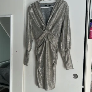 Silver glittrig klänning från Bik Bok - Snygg silverfärgad klänning från Bik Bok med glittrigt material och knytdetalj framtill. Klänningen har långa ärmar och en v-ringad design som ger en elegant look. Perfekt för en kväll ute eller en speciell tillställning.