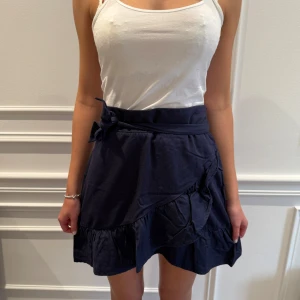 Mörkblå kjol från Holly & Whyte - Snygg mörkblå kjol från Holly & Whyte med volangdetaljer och knytband i midjan. Perfekt för en stilren look. Kjolen har en omlott-design som ger en fin passform.