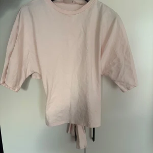 Ljusrosa kortärmad topp med knyt - Säljer en söt ljusrosa topp med korta ärmar och knytning i ryggen. Perfekt för en avslappnad stil. Toppen har en rund halsringning och är gjord i ett mjukt material. Storlek S