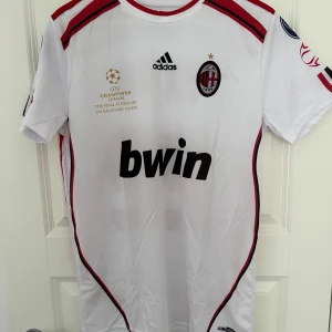 Vit AC Milan fotbollströja med Kaka 22 - Säljer en vit AC Milan fotbollströja från Adidas med Kaka's namn och nummer 22 på ryggen. Tröjan har korta ärmar och röda detaljer samt klubbens emblem på bröstet. Perfekt för fans av laget och spelaren!