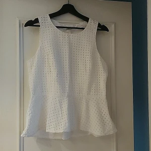 Vit ärmlös topp från H&M - Säljer en stilren vit ärmlös topp från H&M med ett vackert hålmönster. Toppen har en lätt volangdetalj vid nederkanten som ger en feminin touch. Perfekt för varma dagar eller sommarens festligheter.