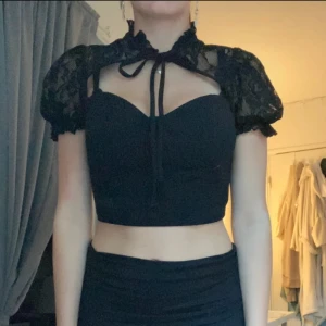 Svart spetsdetaljerad korsettopp - Snygg svart korsettopp med spetsdetaljer och knytning fram. Toppen är croppad och har korta ärmar i spets som ger en elegant touch. Perfekt för en kväll ute eller en speciell tillställning.