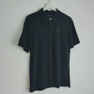 Svart pikétröja från J.Lindeberg - Snygg svart pikétröja från J.Lindeberg i storlek M. Tröjan har en klassisk krage och korta ärmar, perfekt för en stilren look. Tillverkad i Portugal.