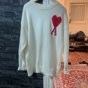 Vit tröja från Ami - Säljer en stilren vit tröja från Ami med ett rött hjärta och bokstaven 'A' broderat på bröstet. Tröjan har långa ärmar och en ribbad rund halsringning. Perfekt för en avslappnad och trendig look.