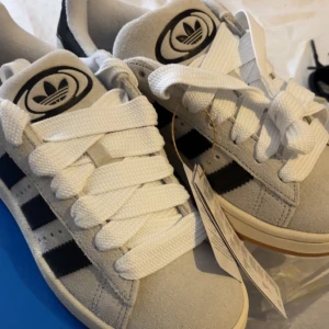 Adidas Campus 00s  - Snygga Adidas Campus 00s W sneakers i grått/vitt och svart med vita detaljer(crystal white core black). Skorna har en klassisk design med snörning och den ikoniska trefoil-loggan på hälen. Perfekta för en stilren och bekväm look. Tillkom även svarta snören om man föredrar det. (Orignalpris 1999kr)