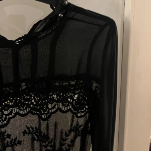 Svart spetsklänning - Elegant svart klänning med vacker spetsdetalj och transparenta partier. Klänningen har långa ärmar och en sofistikerad design som passar perfekt för festliga tillfällen. Passar även som strand klänning. 