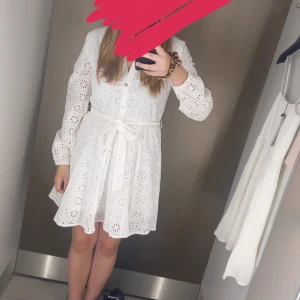 Vit klänning med blomdetaljer - Charmig vit klänning med blomdetaljer och knappar framtill. Den har långa ärmar och ett knytband i midjan för en feminin silhuett. Perfekt för somriga dagar. 