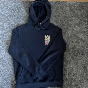 Svart hoodie från Morris - Säljer en stilren svart hoodie från Morris med en broderad logga och brittisk flagga på bröstet. Tröjan har en klassisk känguruficka och justerbar huva med snören. Perfekt för en avslappnad look.