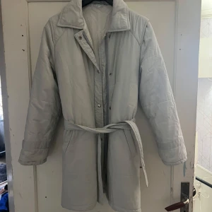 Vit lång kappa med bälte - Säljer en stilren vit lång kappa med bälte i midjan. Jackan har långa ärmar och stängs med knappar. Perfekt för kyligare dagar.