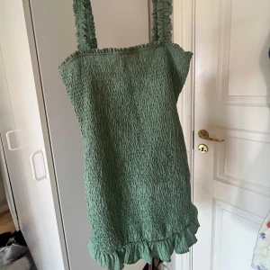 Grön klänning med volangdetaljer - Säljer en söt grön klänning med volangdetaljer i nederkant. Klänningen har breda axelband och en smockad textur som ger en fin passform. Perfekt för sommarens varma dagar!