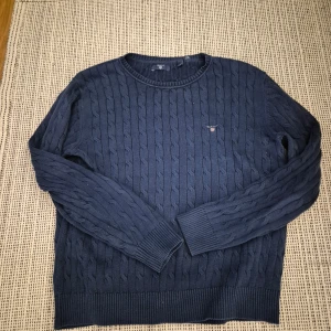 Mörkblå tröja från GANT - Säljer en snygg mörkblå stickad tröja från GANT med kabelstickat mönster. Tröjan har långa ärmar och en rund halsringning. Perfekt för vår dagarna.