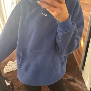 Blå hoodie från Nike - Säljer en snygg blå hoodie från Nike med en liten vit logga på bröstet. Den har en klassisk känguruficka och ribbade muddar vid ärmslut. Perfekt för en avslappnad stil! 💙