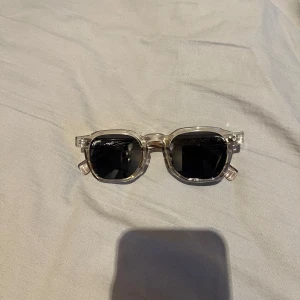 Transparenta solglasögon med svart lins - Snygga solglasögon med transparent båge och svarta linser. Bågarna har en unik, kantig design och detaljer på skalmarna som ger en extra touch. Perfekta för en stilren look.