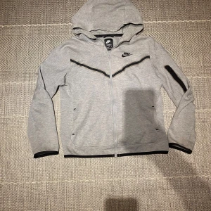 Grå huvtröja från Nike - Säljer en grå huvtröja från Nike med dragkedja och huva. Tröjan har svarta detaljer och en ficka med dragkedja på vänstra armen. Pris kan diskuteras vid snabb affär. Skick: 8/10