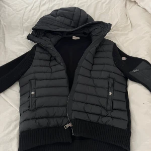 Svart jacka från Moncler - Säljer en snygg svart dunväst från Moncler med huva och dragkedja. Västen har ribbade detaljer och två fickor med knappar framtill. Perfekt för kyligare dagar.