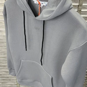 Grå hoodie från Off-White - Snygg grå hoodie från Off-White med svart dragsko och stor ficka framtill. Baksidan pryds av det ikoniska Off-White-mönstret i ljusare grått. Perfekt för en stilren och avslappnad look.