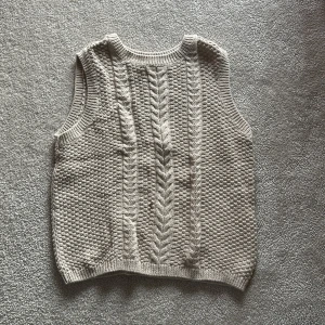 Beige stickad väst från H&M - Säljer en beige stickad väst från H&M i storlek 140. Västen har ett snyggt flätmönster framtill och är ribbad vid ärmhål och nederkant. Perfekt för lager-på-lager under kyligare dagar.