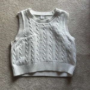 Vit stickad väst från Lindex - Säljer en snygg vit stickad väst från Lindex. Västen har ett klassiskt flätmönster och ribbade kanter. Perfekt för lager-på-lager-stil under svalare dagar. Tillverkad i en mjuk bomullsblandning.