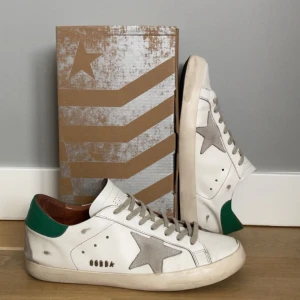Golden Goose Super-Star sneakers - Snygga vita sneakers från Golden Goose med grön häl och grå stjärndetalj på sidan. Skorna har snörning och GGDB-logga. Perfekta för en avslappnad stil. Väldigt bra skick , storlek 44. Några synliga defekter men annars som gått som nya, därför den med sätta priset. Hör av dig vid minsta fundering. Pris kan självklart diskuteras.