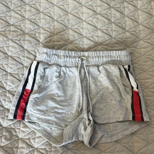 Grå shorts från Tommy Hilfiger - Snygga grå shorts från Tommy Hilfiger med elastisk midja och snörning. De har coola ränder i svart, vitt och rött längs sidorna. Perfekta för en avslappnad stil.