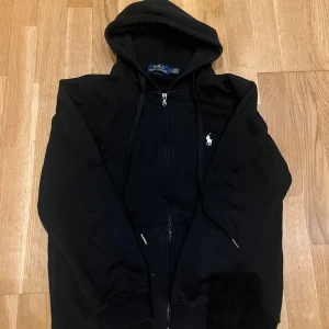 Svart hoodie från Ralph Lauren - Säljer en stilren svart hoodie från Ralph Lauren med dragkedja och klassisk logga på bröstet. Perfekt för en avslappnad look. Den har en justerbar huva och ribbade muddar för extra komfort.