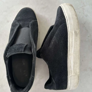 Svarta sneakers från Axel Arigato - Snygga svarta sneakers från Axel Arigato med en stilren design. Skorna har en elastisk rem över foten istället för traditionell snörning och en vit sula som ger en fräsch look. Man får även med låda och stickers. 38,5 men passar 38-39 priset är ej hugget i sten