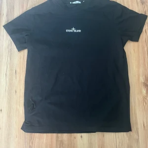 Svart t-shirt från Stone Island - Säljer en svart t-shirt från Stone Island med en liten logga på bröstet och en stor grafisk design på ryggen. Perfekt för en stilren och modern look. T-shirten är kortärmad och gjord i bomull. (Pris kan diskuteras vid snabb affär)