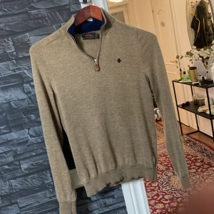 Beige tröja med dragkedja från Morris - Säljer en stilren beige tröja från Morris med dragkedja vid halsen. Tröjan har en liten broderad detalj på bröstet och är tillverkad i mjukt material. Perfekt för en avslappnad men snygg look.