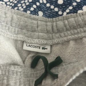 Grå mjukisbyxor från Lacoste - Säljer ett par grå mjukisbyxor från Lacoste med elastisk midja och grön snörning. Byxorna har det klassiska Lacoste-logotypen broderad på sidan. Perfekta för en avslappnad stil.
