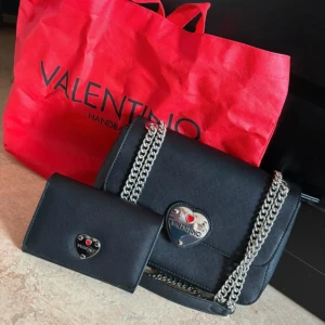 Svart axelväska och plånbok från Valentino - Snygg svart axelväska och matchande plånbok från Valentino. Väskan har en kedjerem och ett hjärtformat emblem med märkeslogga. Perfekt för att ge en stilren touch till din outfit.