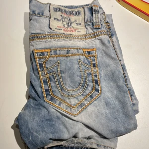 Ljusblå jeans från True Religion - Säljer ett par ljusblå jeans från True Religion med slitna detaljer och kontrastsömmar. Jeansen har en straight passform och klassiska femficksdesign. Perfekta för en avslappnad stil.