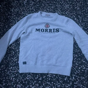 Grå tröja från Morris - Säljer en stilren grå tröja från Morris med text och emblem på bröstet. Tröjan har långa ärmar och ribbade kanter vid hals, ärmslut och nederkant. Perfekt för en klassisk look.