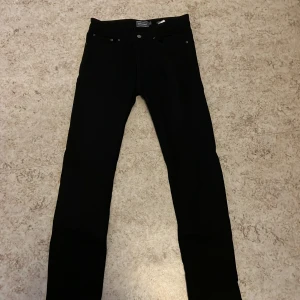 Svarta jeansbyxor från Dressmann - Säljer ett par svarta jeansbyxor från Dressmann i regular fit. Byxorna har en klassisk design med knappar och dragkedja framtill. Perfekta för en stilren look.