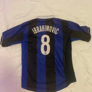 Ibrahimovic tröja - Säljer en klassisk blå och svart randig fotbollströja från Nike med spelarnamnet Ibrahimovic och nummer 8 på ryggen. Tröjan har korta ärmar och klubbens emblem på bröstet. Perfekt för fotbollsfans!