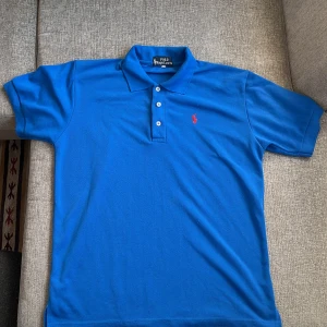 Blå pikétröja från Ralph Lauren - Säljer en klassisk blå pikétröja från Ralph Lauren med röd logga på bröstet. Tröjan har korta ärmar och tre knappar vid kragen. 