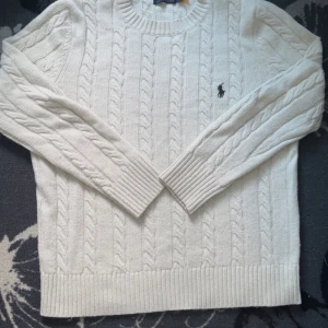 Vit stickad tröja från Ralph Lauren - Säljer en klassisk vit stickad tröja från Ralph Lauren med kabelstickat mönster och broderad logga på bröstet. Tröjan har långa ärmar och ribbstickade muddar vid ärmslut och nederkant. Perfekt för kyliga dagar!