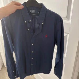 Blå skjorta från Polo Ralph Lauren - Snygg blå skjorta från Polo Ralph Lauren med klassisk krage och knappar framtill. Den har en broderad röd logotyp på bröstet och långa ärmar. Perfekt för en stilren look.