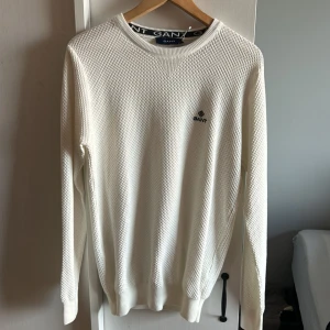 Vit tröja från GANT - Säljer en stilren vit tröja från GANT med ett diskret mönster och logotyp på bröstet. Tröjan har långa ärmar och rund halsringning, perfekt för en klassisk look.