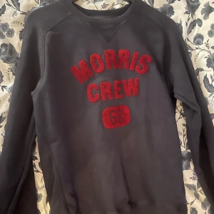 Svart tröja från Morris Gents - Säljer en svart tröja från Morris Gents med röd text 'Morris Crew 68' på framsidan. Tröjan har långa ärmar och en klassisk rund halsringning. Perfekt för en avslappnad stil.