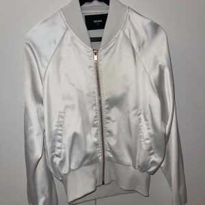 Vit satin bomberjacka från BikBok - Snygg vit bomberjacka från BikBok i satinliknande material. Jackan har en glansig finish, ribbade muddar och dragkedja i roséguld. Klassisk bombermodell med fickor på sidorna och lång ärm. Perfekt för dig som vill sticka ut med en clean look.