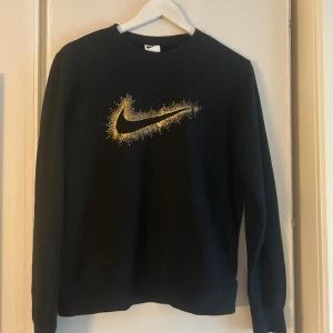 Svart Nike tröja med guldlogo - Svart långärmad tröja från Nike med en stor guldig swoosh-logga framtill som har coolt stänkmönster. Klassisk rund halsringning och mjukt material, perfekt för dig som gillar sportig och stilren vibe.