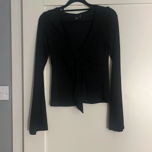 Svart långärmad topp med knyt Gina Tricot - Svart långärmad topp från Gina Tricot i storlek XS. Djup v-ringning med snygg knytning framtill som ger en trendig look. Tunn och stretchig i materialet, perfekt till lager-på-lager eller som statement-piece.