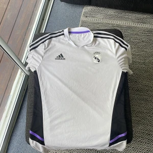 Real Madrid Adidas fotbollströja vit - Snygg Real Madrid tränings tröja ifrån 2022/23 säsongen. Den är  från Adidas i vitt med svarta detaljer och lila inslag. Tröjan har korta ärmar, klassiska Adidas-ränder på axlarna och klubbens emblem på bröstet. Tillverkad i lätt och ventilerande material, perfekt för träning eller match.