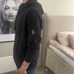 Svart hoodie från Stone Island - Svart hoodie från Stone Island med klassisk patch på vänster ärm. Tröjan har huva med snörning, magficka och ribbade muddar. Perfekt för dig som gillar streetwear och vill ha en clean look med ikonisk detalj.
