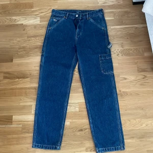 Tommy Jeans blå cargopants - Snygga blå cargopants från Tommy Jeans med raka ben och flera praktiska fickor på sidorna. Klassisk jeanslook med tydliga sömmar och logga på bakfickan. Tillverkade i robust denim som ger en cool och avslappnad vibe.