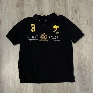 Svart pikétröja från Vinson Polo Club - Säljer en svart pikétröja från Vinson Polo Club med broderade detaljer i gult och vitt framtill. Tröjan har klassisk krage, korta ärmar och tryck med klubbens logga samt siffran 3 på bröstet.