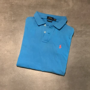 Blå pikétröja från Ralph Lauren - Säljer en klassisk blå pikétröja från Ralph Lauren, modell The Skinny Polo. Tröjan har korta ärmar, krage och knäppning framtill samt den ikoniska röda loggan broderad på bröstet. Tröjan är i storlek L för barn. Tröjan har också några edfekter som att den är blekt, ni ser på bild 4 och 5. Bara att höra av sig vid funderingar eller frågor! Priset kan diskuteras!😀